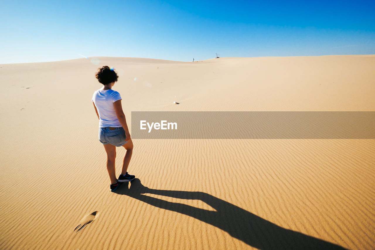 Rear view of woman walking on sand against clear sky at desert during sunny day