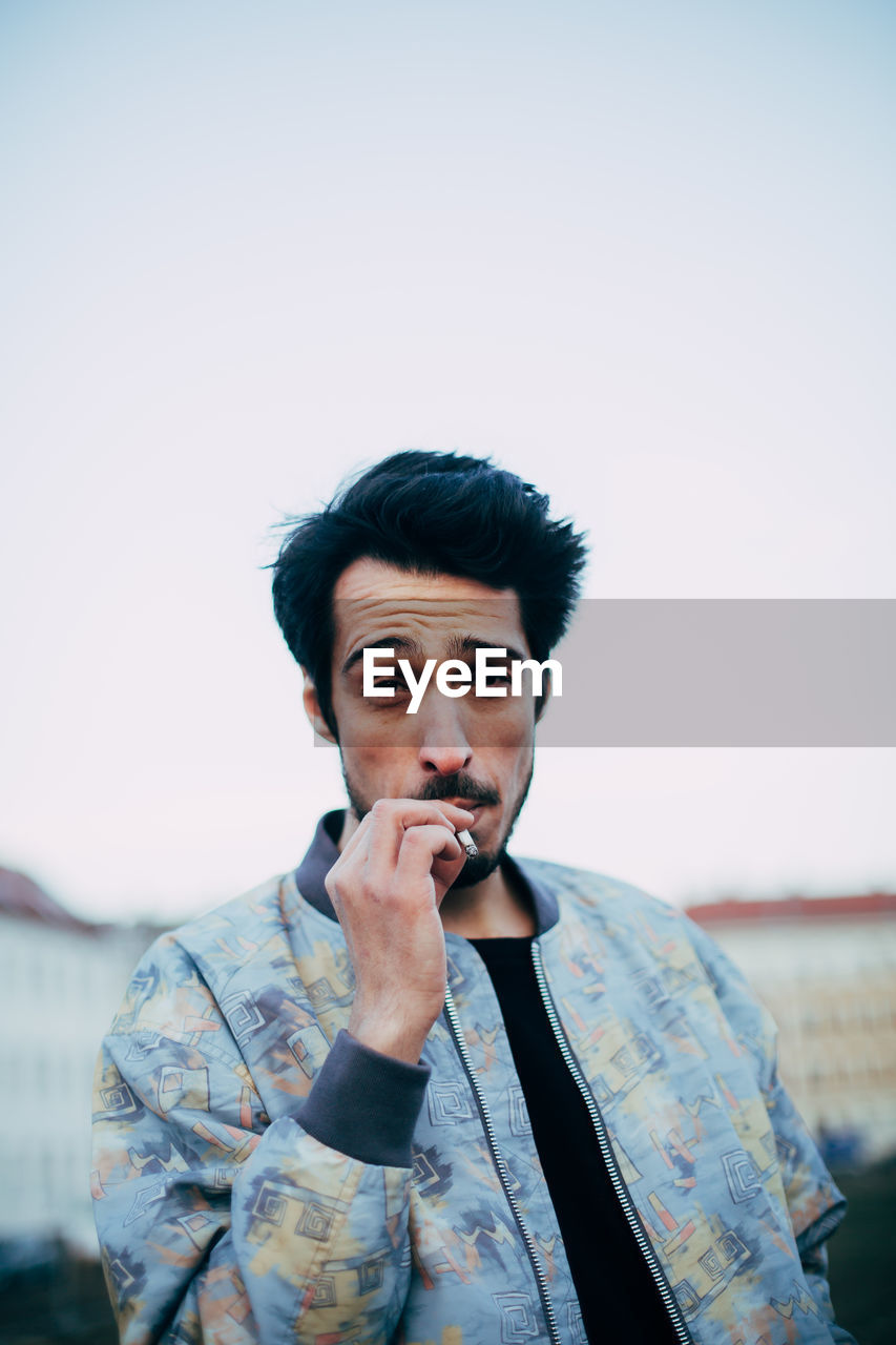 Portrait of young man smoking cigarette against sky