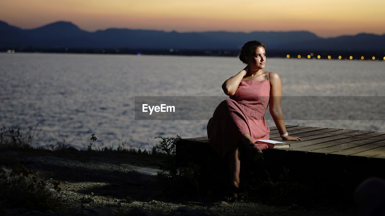 Portrait of woman sitting on shore against sky during sunset