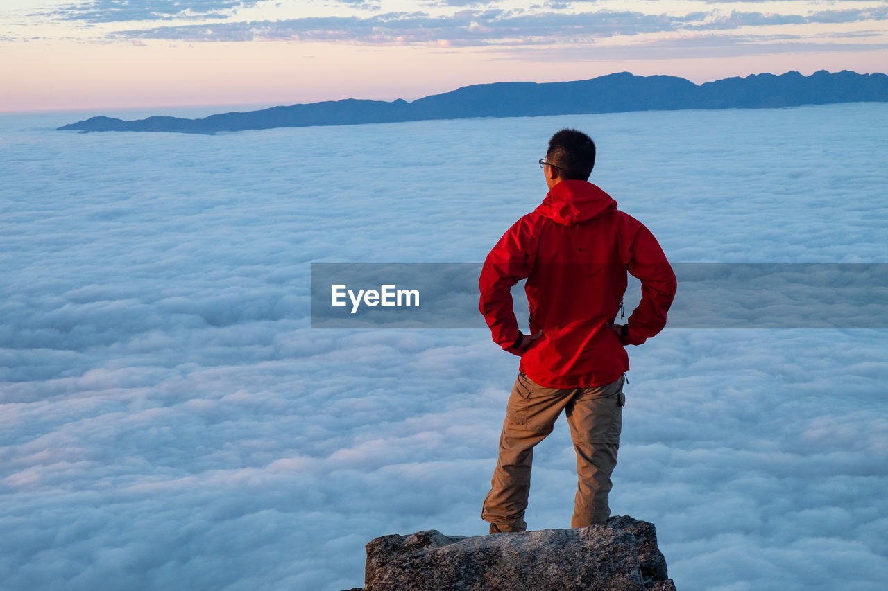 Man in red jacket  standing and looking at the clouds below 