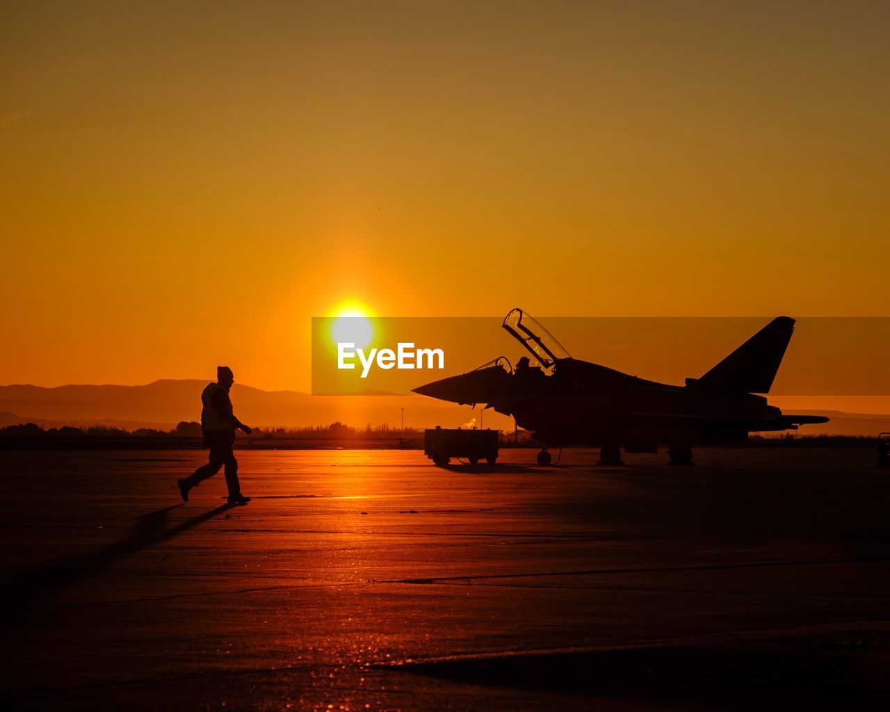 Silhouette man walking by fighter plane on runway during sunset