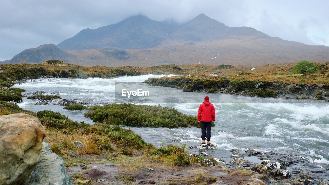 Rear view of person standing by flowing river against mountains