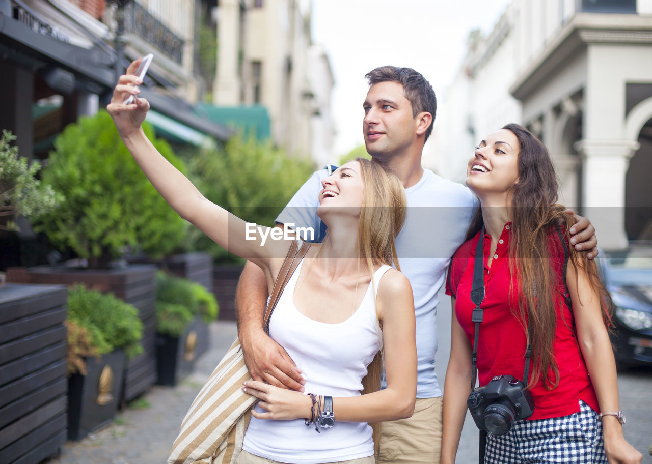 HAPPY YOUNG WOMAN PHOTOGRAPHING WHILE STANDING IN CITY