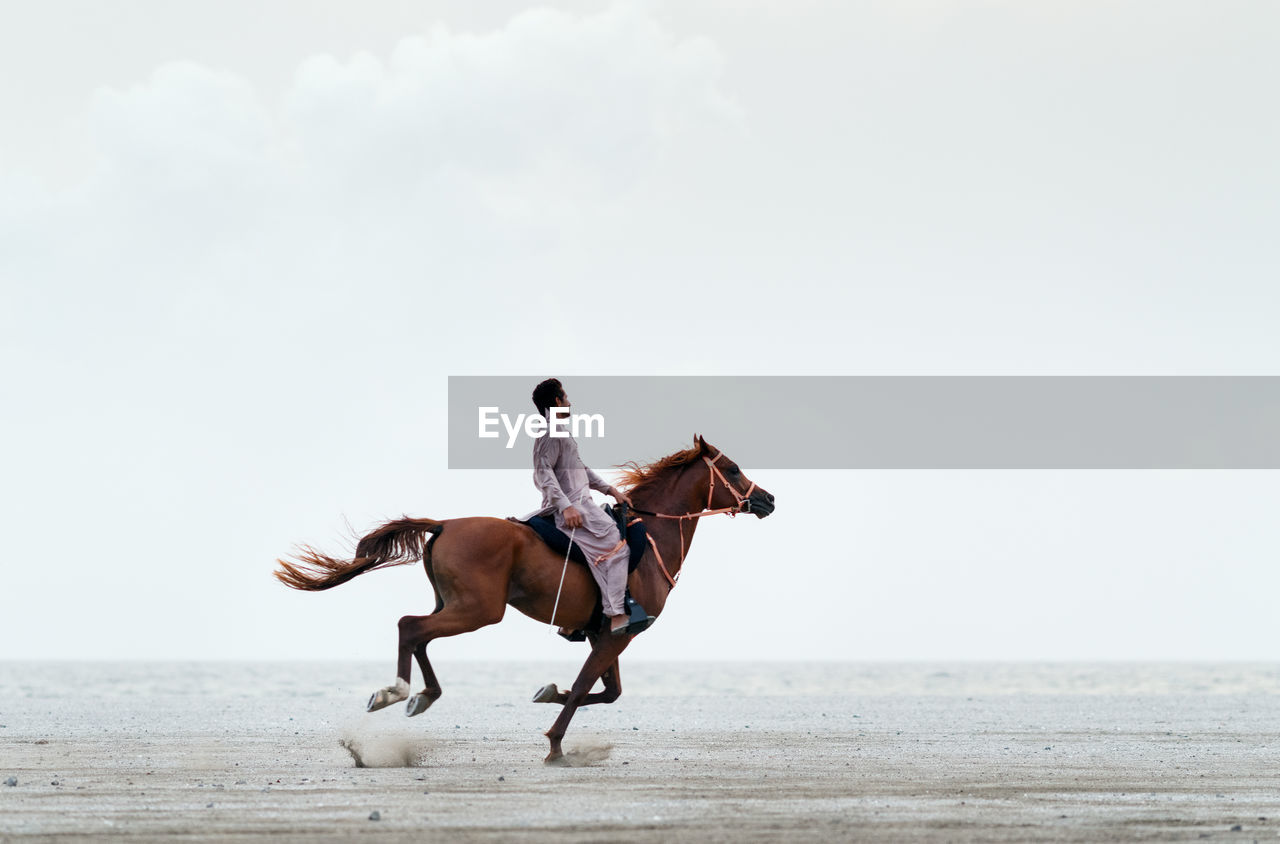 Side view full length of man riding horse on sand against clear sky