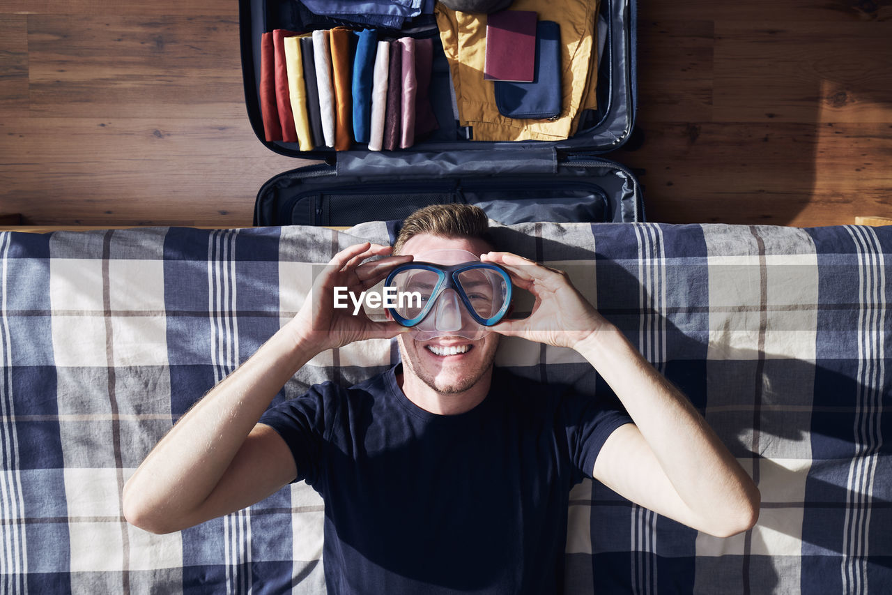 Funny portrait of happy young man during packing suitcase and planning travel adventure.