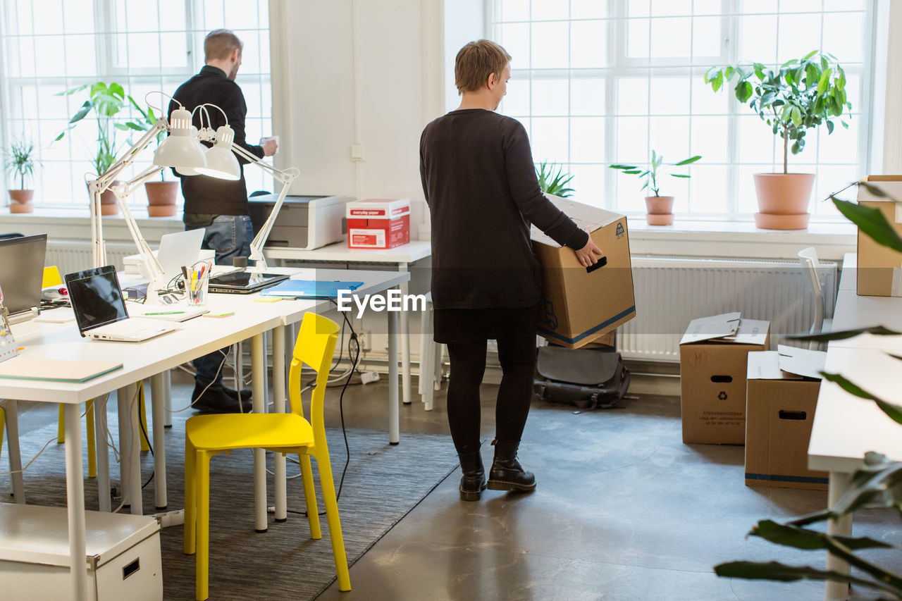 Rear view of businesswoman carrying cardboard box by desk in creative office