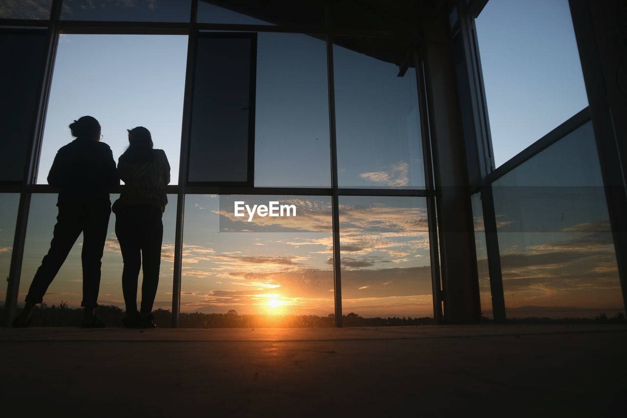 Low angle view silhouette of people standing by window against sky during sunset