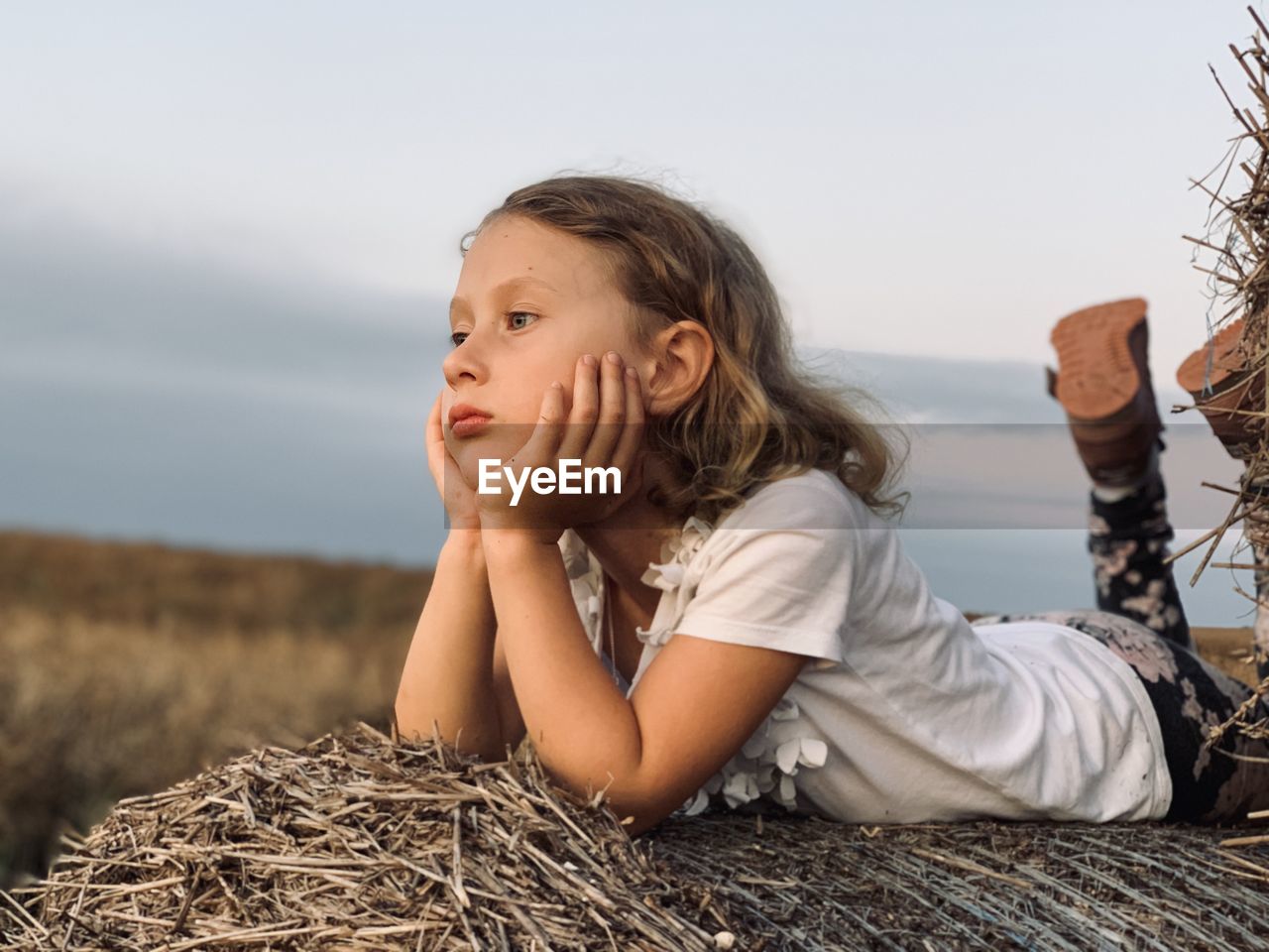 Girl looking away while lying on hay against sky during sunset