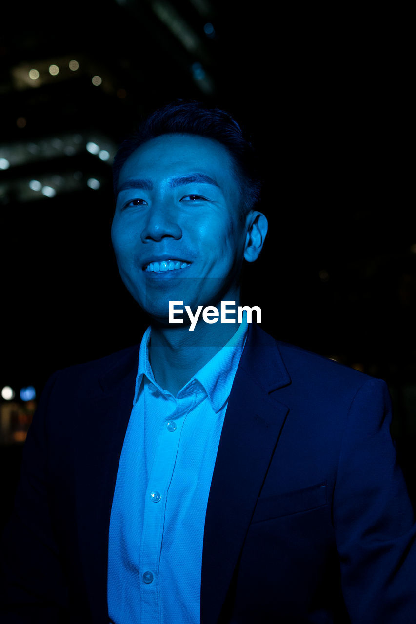 Positive asian male in formal wear looking at camera on street in night time with blue neon light