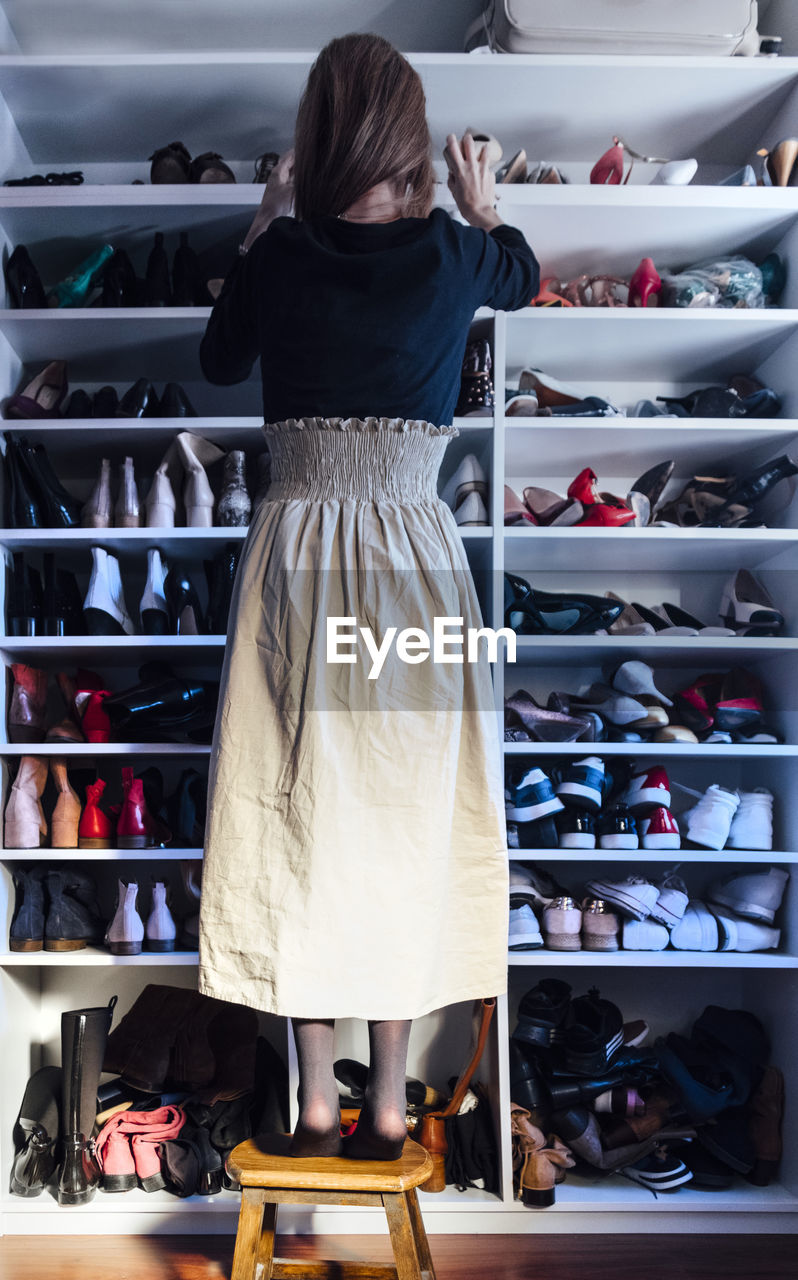 Back view of faceless woman standing on a wooden stool taking beige high heels shoes from shelf of modern white closet