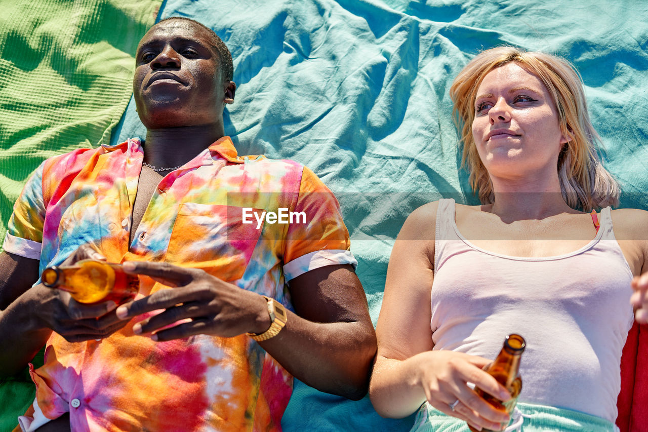 Top view of relaxed young multiracial couple in casual clothes lying on blanket with bottles of beer and looking away during summer holidays