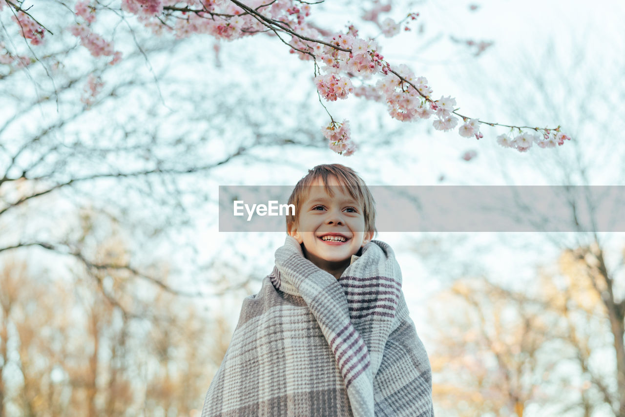 Portrait of a little boy wrapped in a blanket enjoying cherry blossoms in a city park.