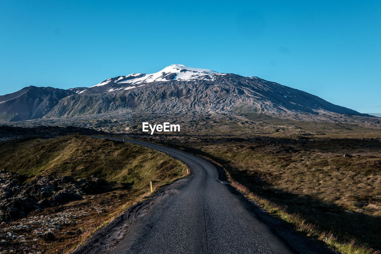 Road amidst snowcapped mountains against clear blue sky