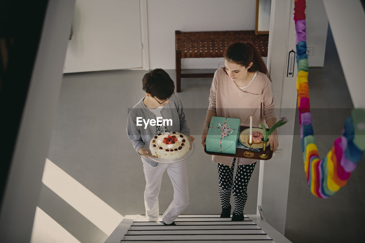 High angle view of siblings carrying birthday cake and gift box while climbing up steps at home