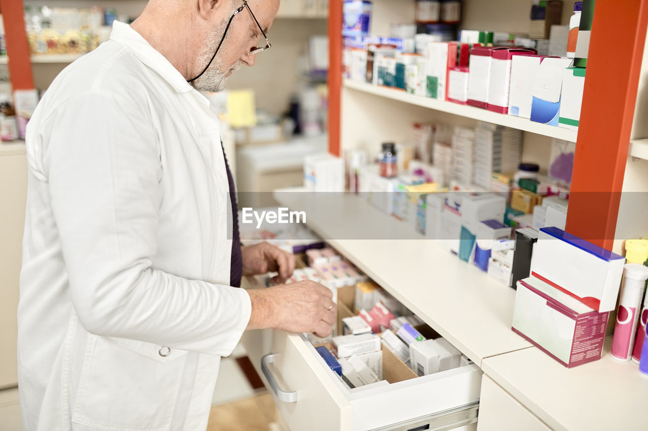 Pharmacist wearing eyeglasses opening drawer of medicines at pharmacy store