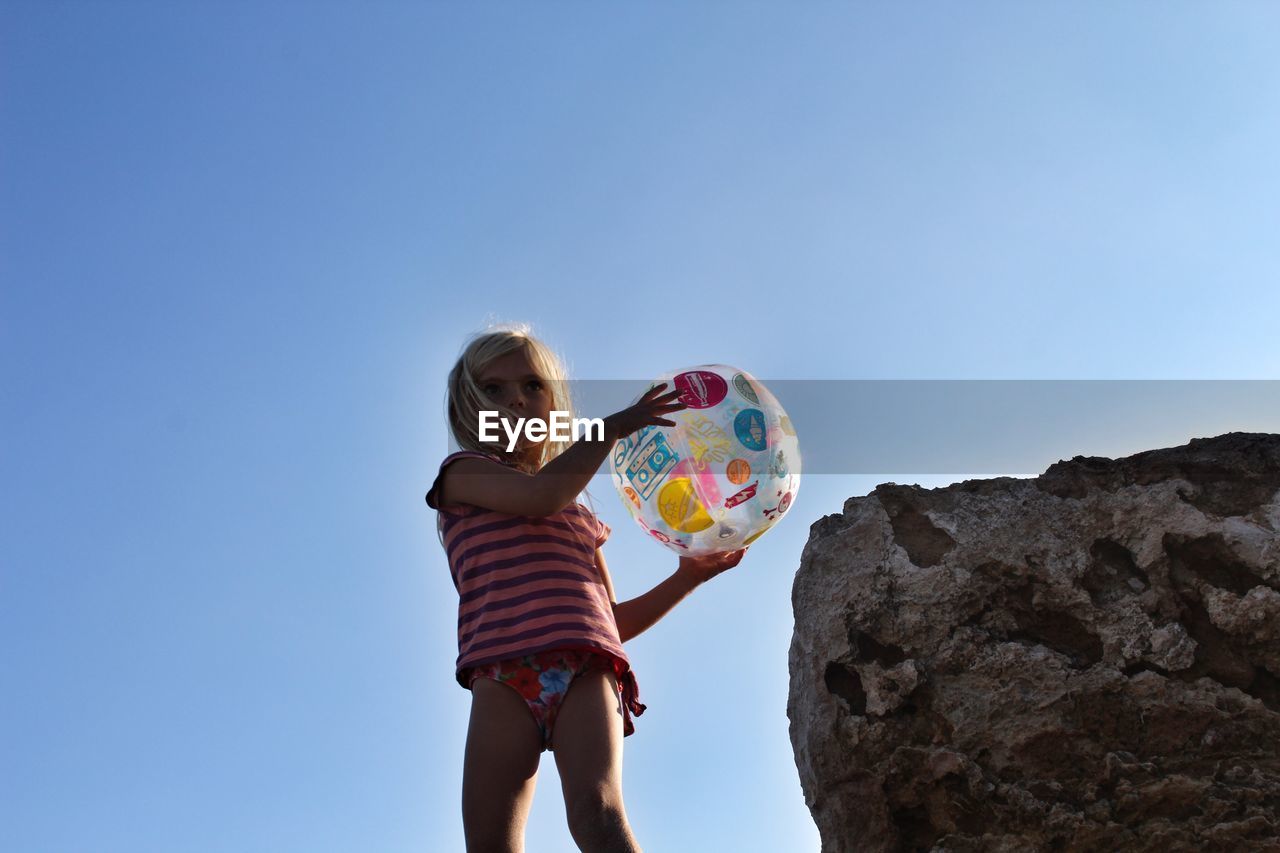 Low angle view of girl holding beach ball while standing by rock against clear sky