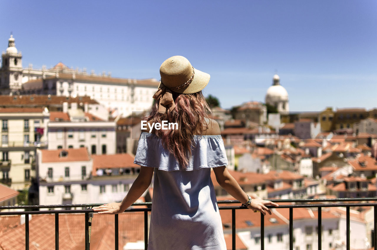 Rear view of young woman looking at buildings while standing by railing in balcony against clear sky