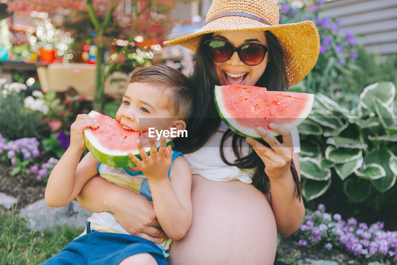 Pregnant mother and son eating watermelons while sitting against plants