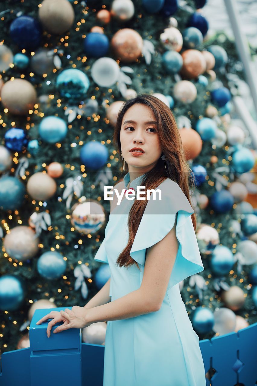 Smiling young woman looking away while standing by christmas tree outdoors