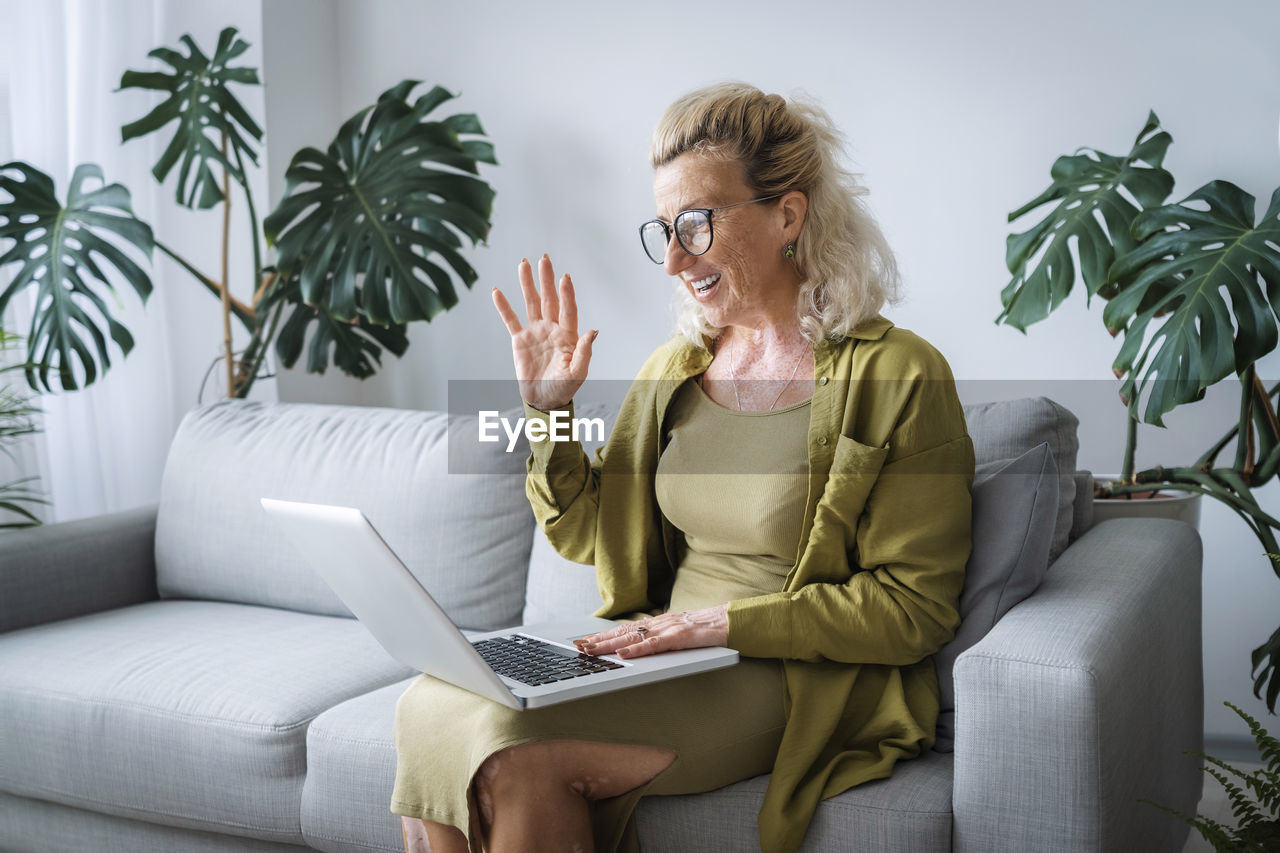 Senior freelancer waving on video call through laptop sitting at home