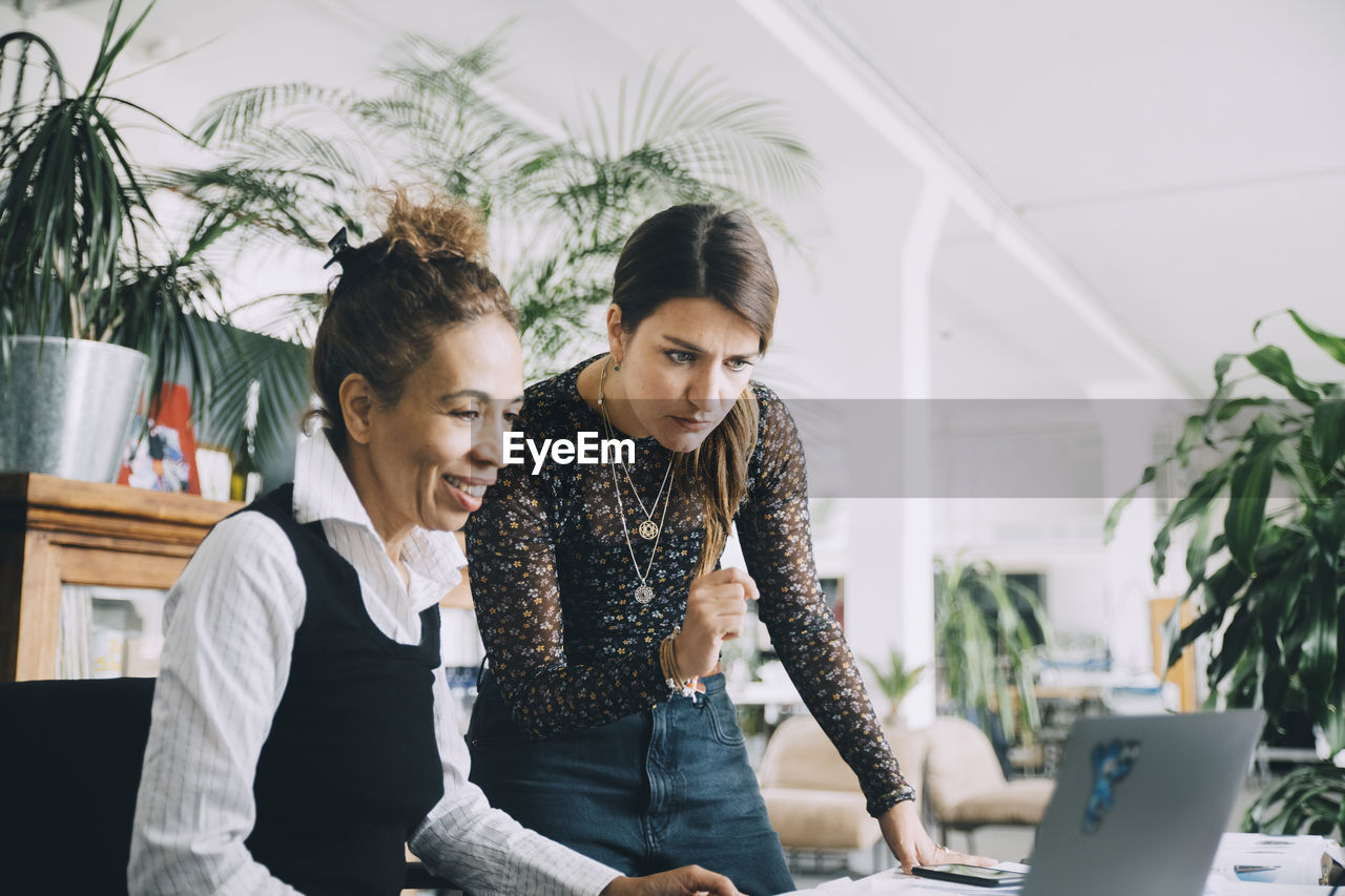 Businesswoman looking while smiling female colleague using laptop in creative office