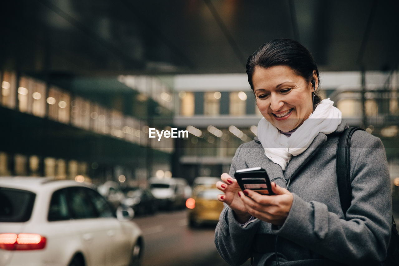 Smiling businesswoman using smart phone while standing on city street against building