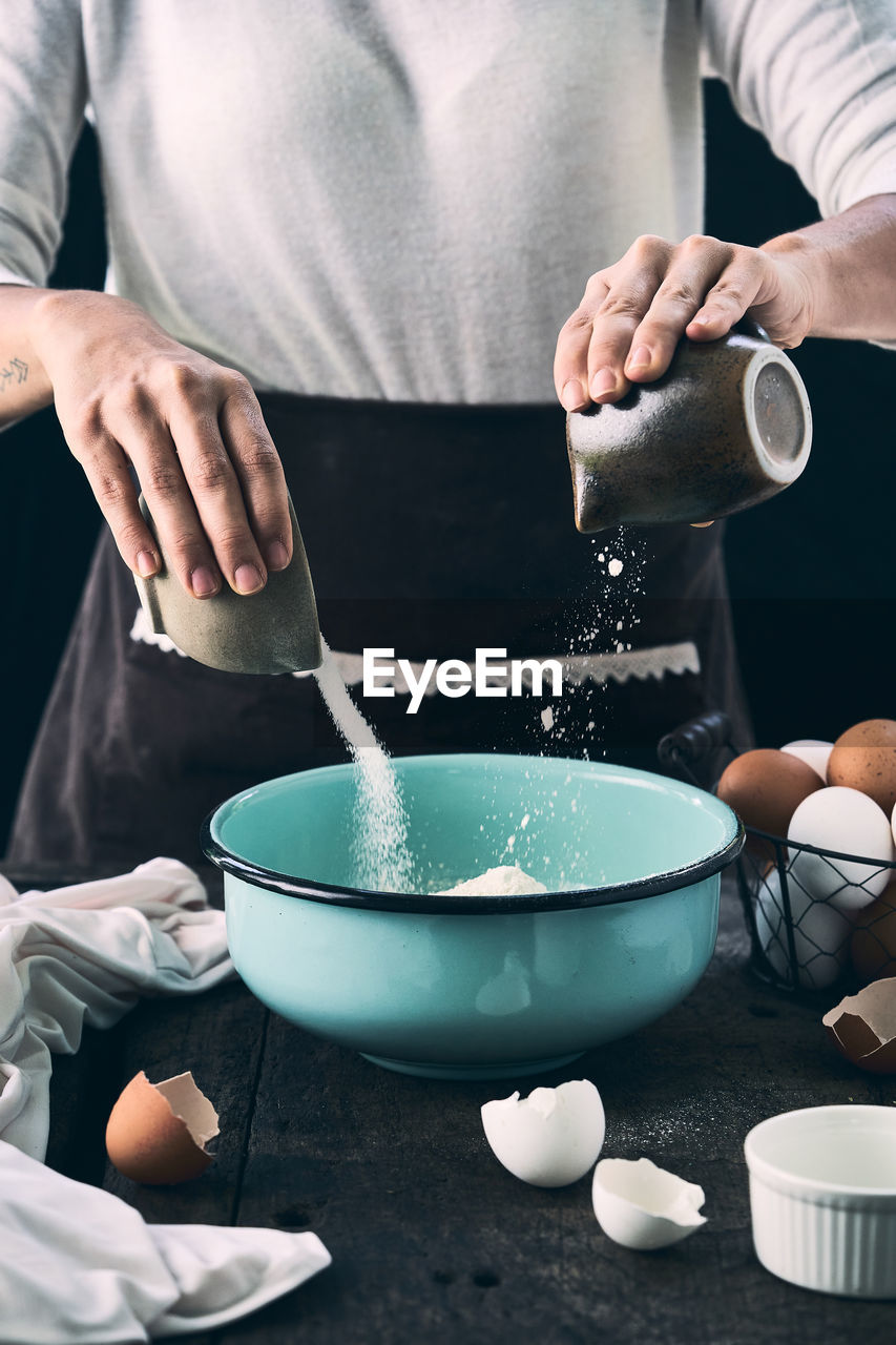 Unrecognizable female cook in apron adding sugar and flour in bowl while mixing ingredients for cooking dough at table