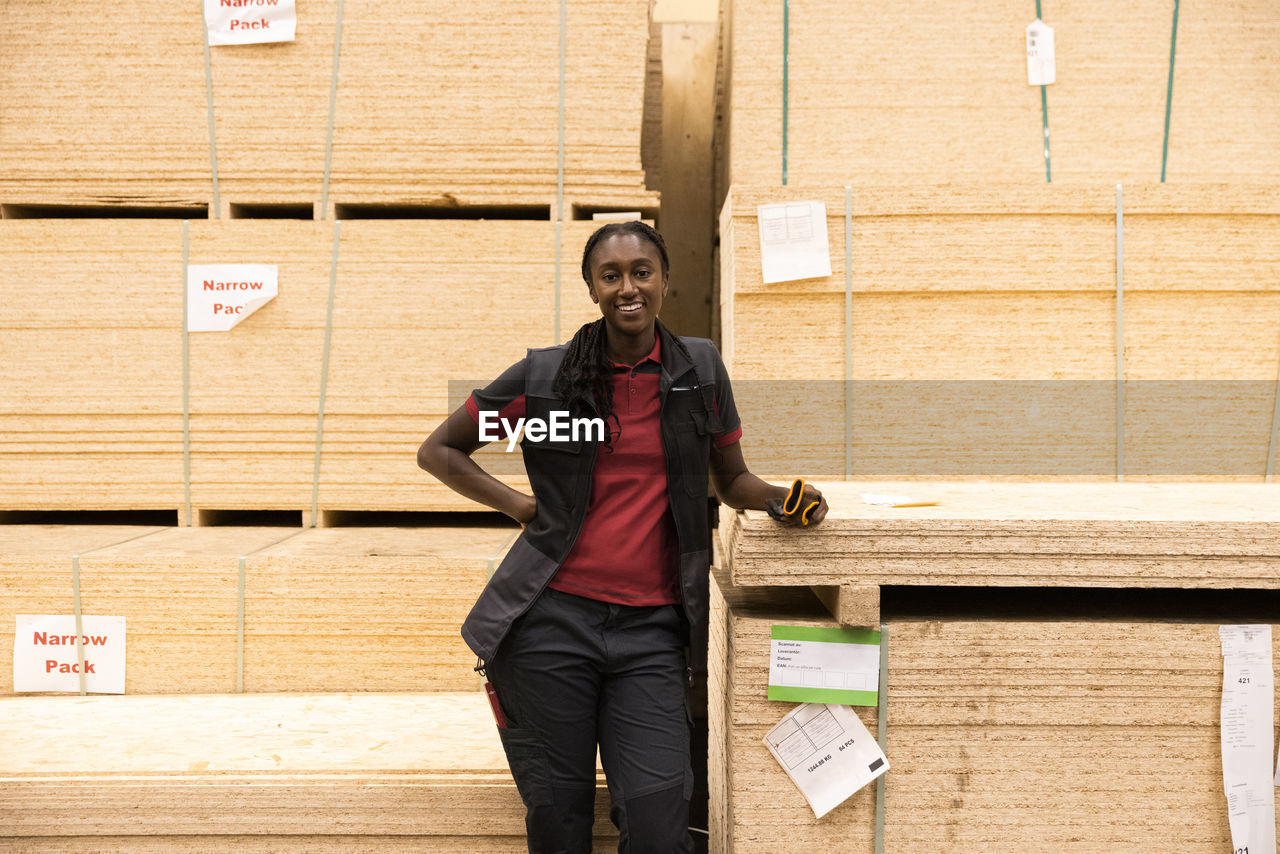 Portrait of smiling saleswoman with hand on hip standing near plywood stack at hardware store