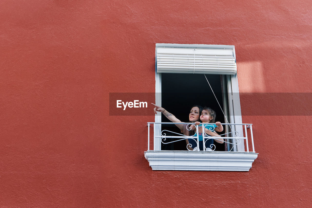 Woman clapping in the window of her house with her daughter on a red background