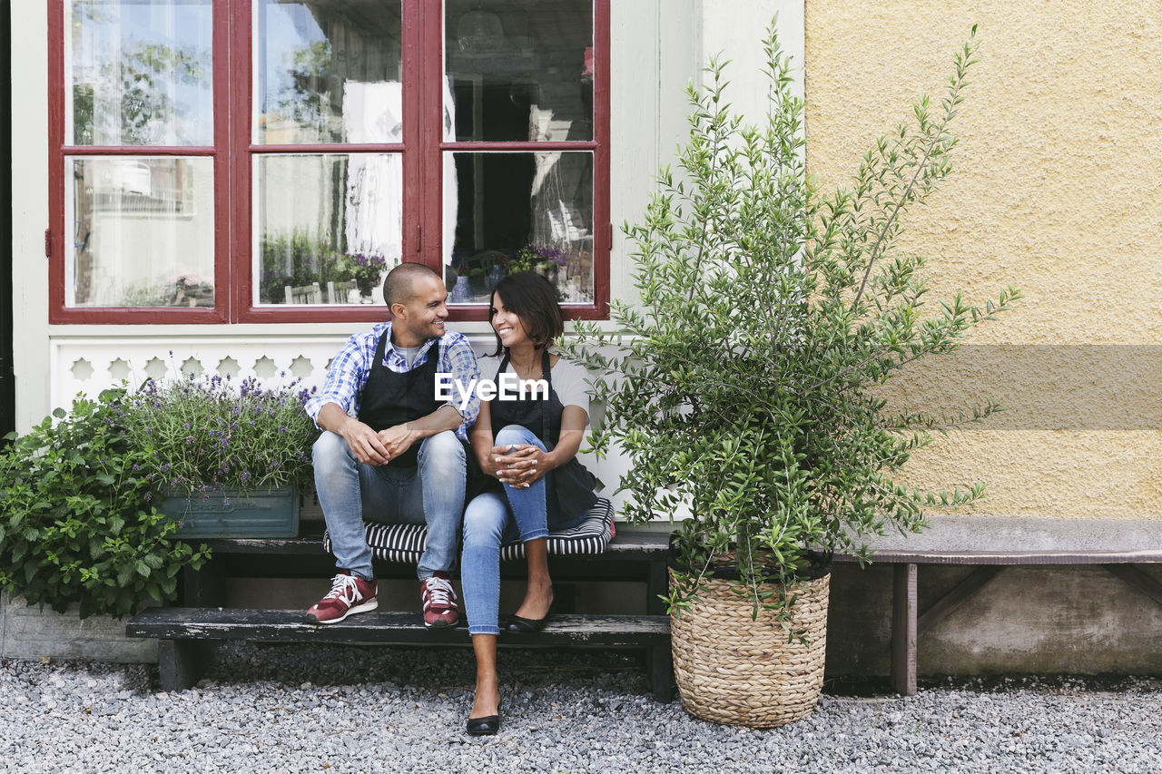 Smiling owners sitting on bench against window outside cafe