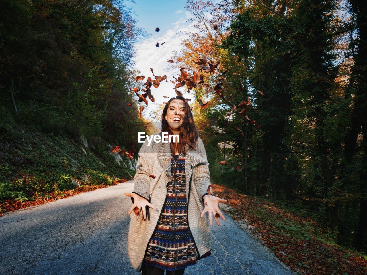 Cheerful woman throwing leaves while standing on road amidst trees