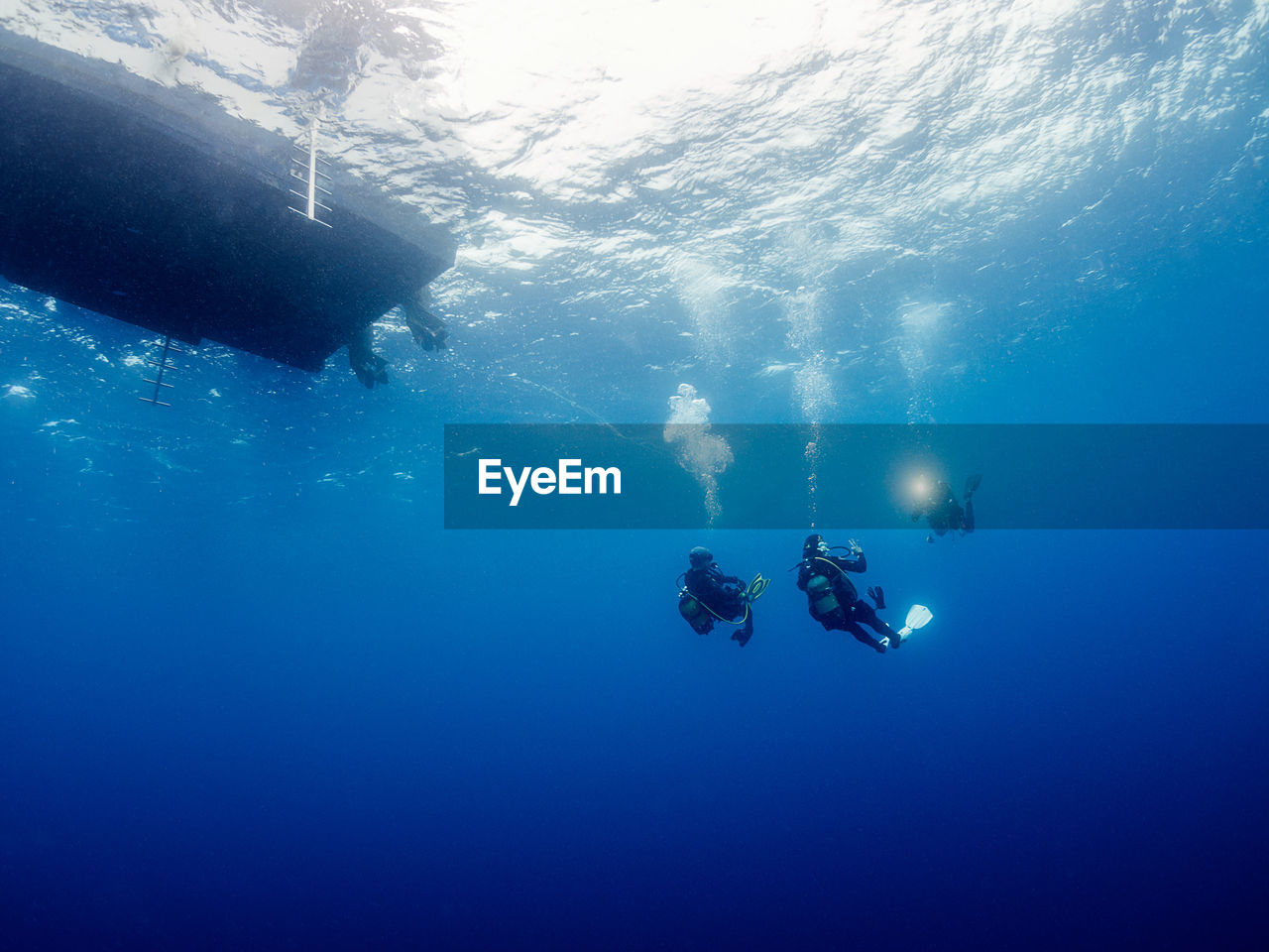 Group of professional divers in wetsuits taking picture of each other while swimming near water surface with boat undersea in deep sea