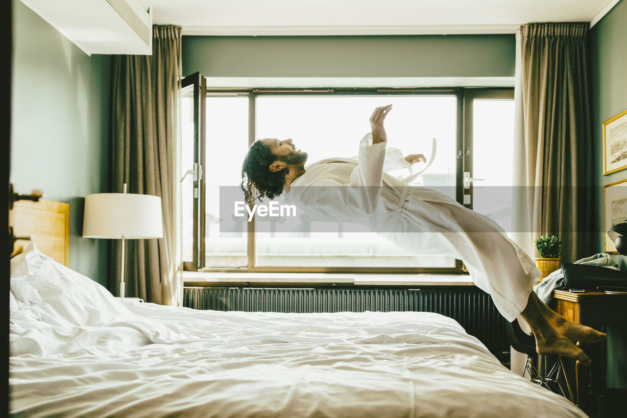 Playful man wearing robe jumping on bed at hotel