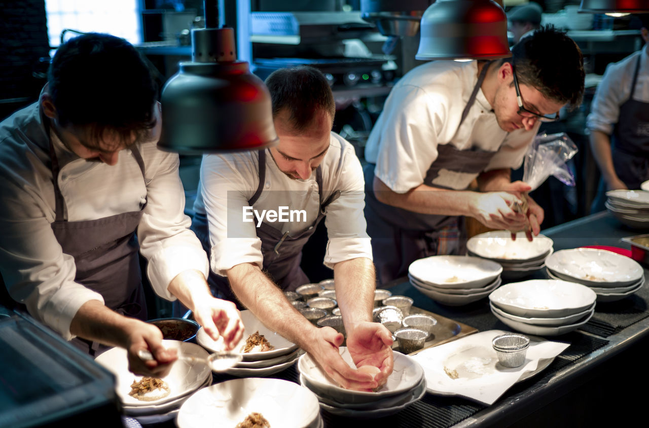 Young cooks men in aprons carefully serving meal in white ceramic dishes on restaurant kitchen