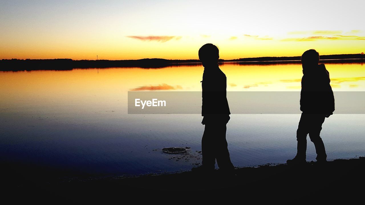 SILHOUETTE MEN STANDING ON LAKE AGAINST SKY DURING SUNSET