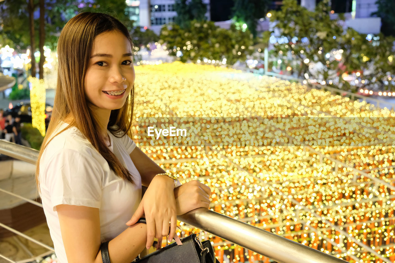 Portrait of smiling young woman standing by railing over illuminated lighting equipment