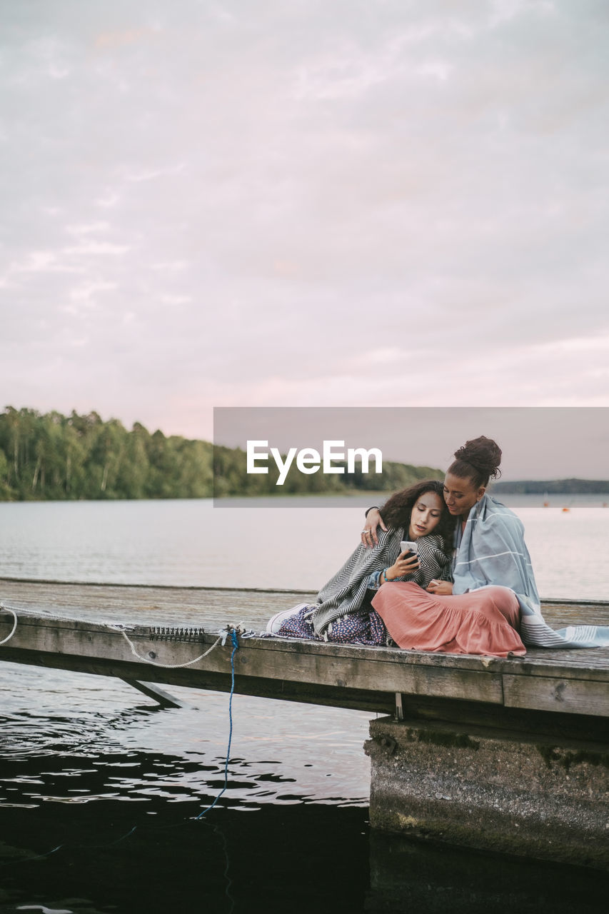 Daughter showing smart phone to mother while sitting on pier over lake during sunset