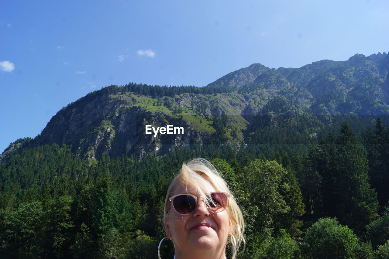 sunglasses, mountain, glasses, tree, one person, plant, portrait, nature, fashion, leisure activity, sky, adult, headshot, mountain range, women, beauty in nature, scenics - nature, lifestyles, land, environment, day, vacation, forest, outdoors, blond hair, travel, trip, adventure, green, holiday, landscape, smiling, sunlight, front view, wilderness, walking, tourism, happiness, young adult, female, travel destinations, blue, summer, pine tree, tranquility, tourist, goggles