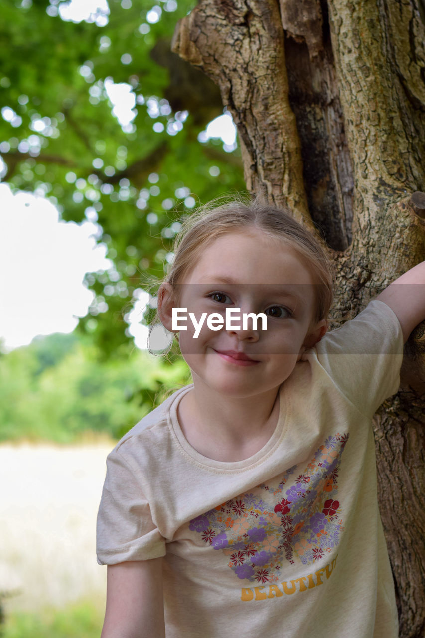 childhood, child, portrait, tree, spring, smiling, plant, looking at camera, female, one person, women, front view, emotion, happiness, portrait photography, tree trunk, trunk, nature, person, day, casual clothing, innocence, cute, blond hair, leisure activity, outdoors, toddler, flower, waist up, standing, cheerful, focus on foreground, skin, enjoyment, headshot, human face, clothing, fun, lifestyles, green, positive emotion, summer, hairstyle, carefree