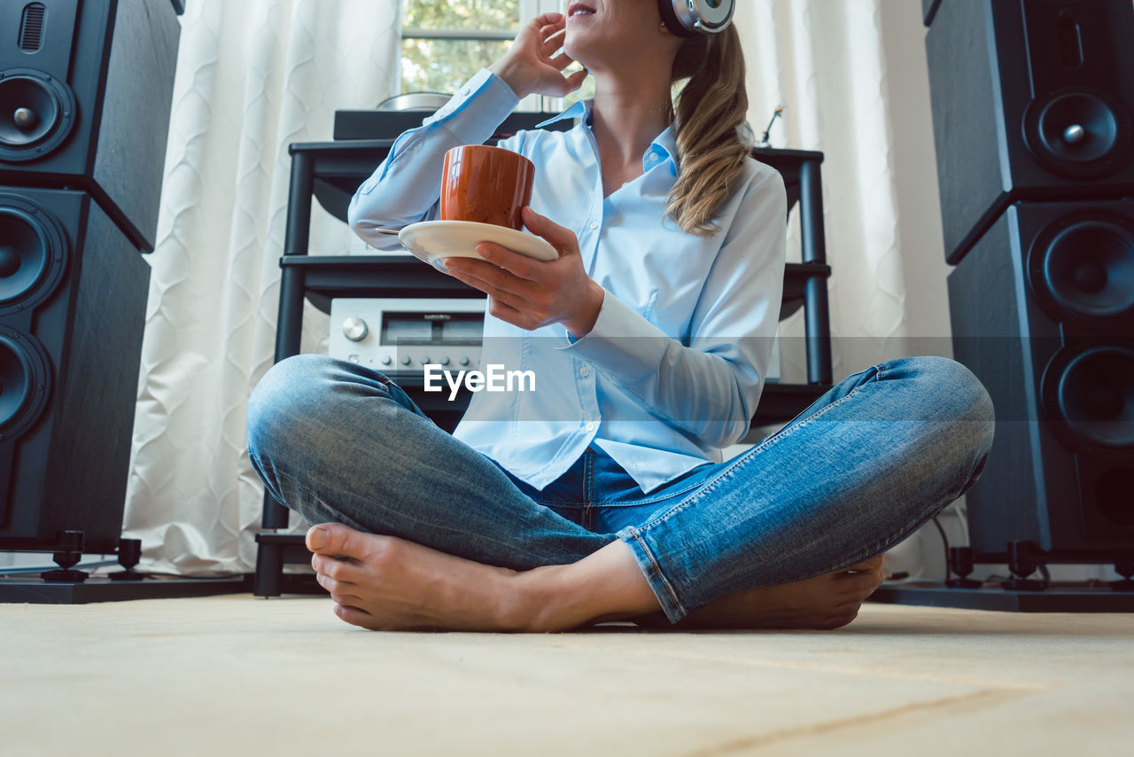 Low section of woman listening music while holding coffee cup on floor