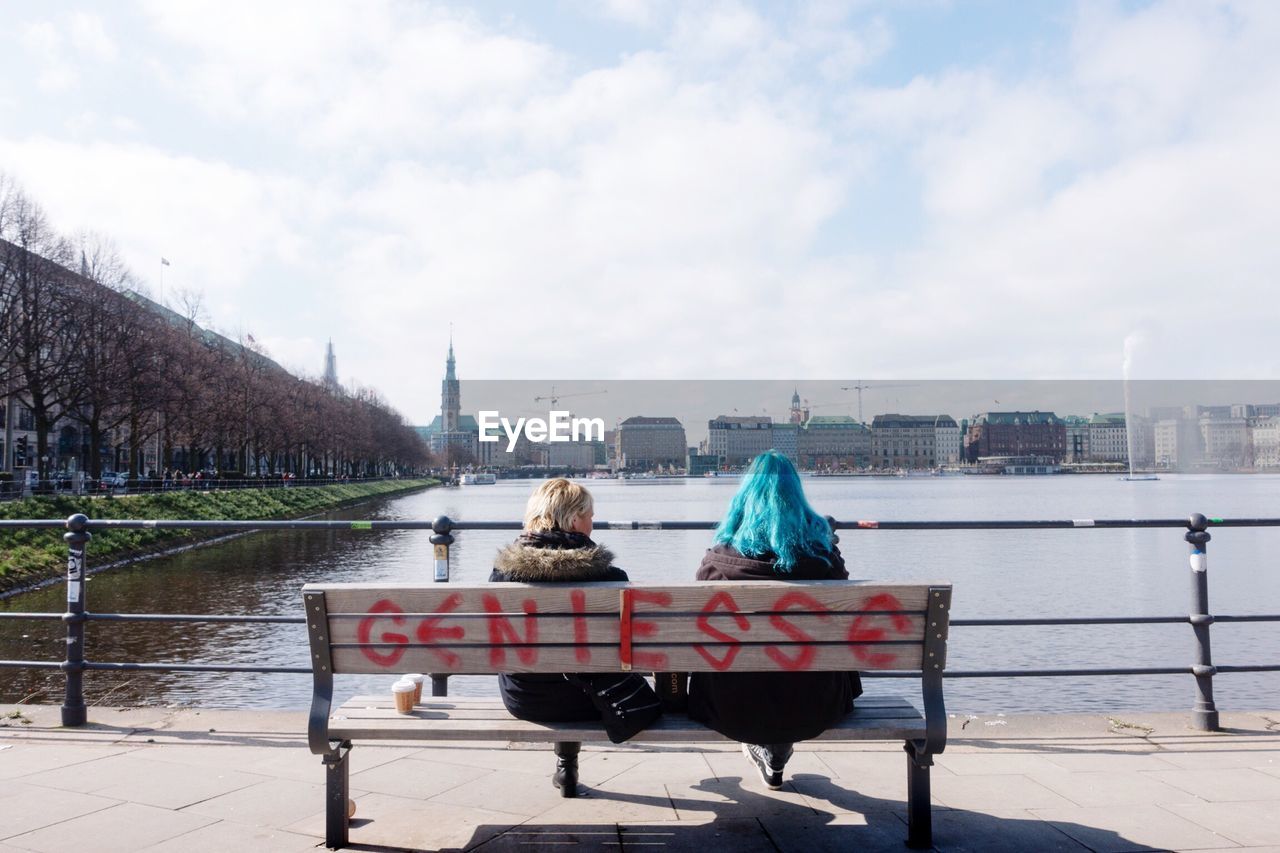 Rear view of women sitting on bench at alster river against sky