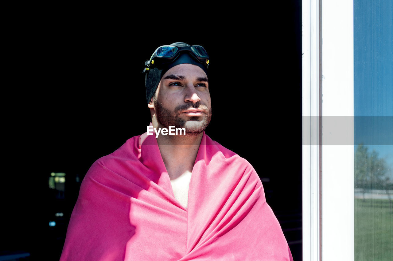 Through window view of dreamy male athlete in swimming cap and goggles with towel looking away on black background
