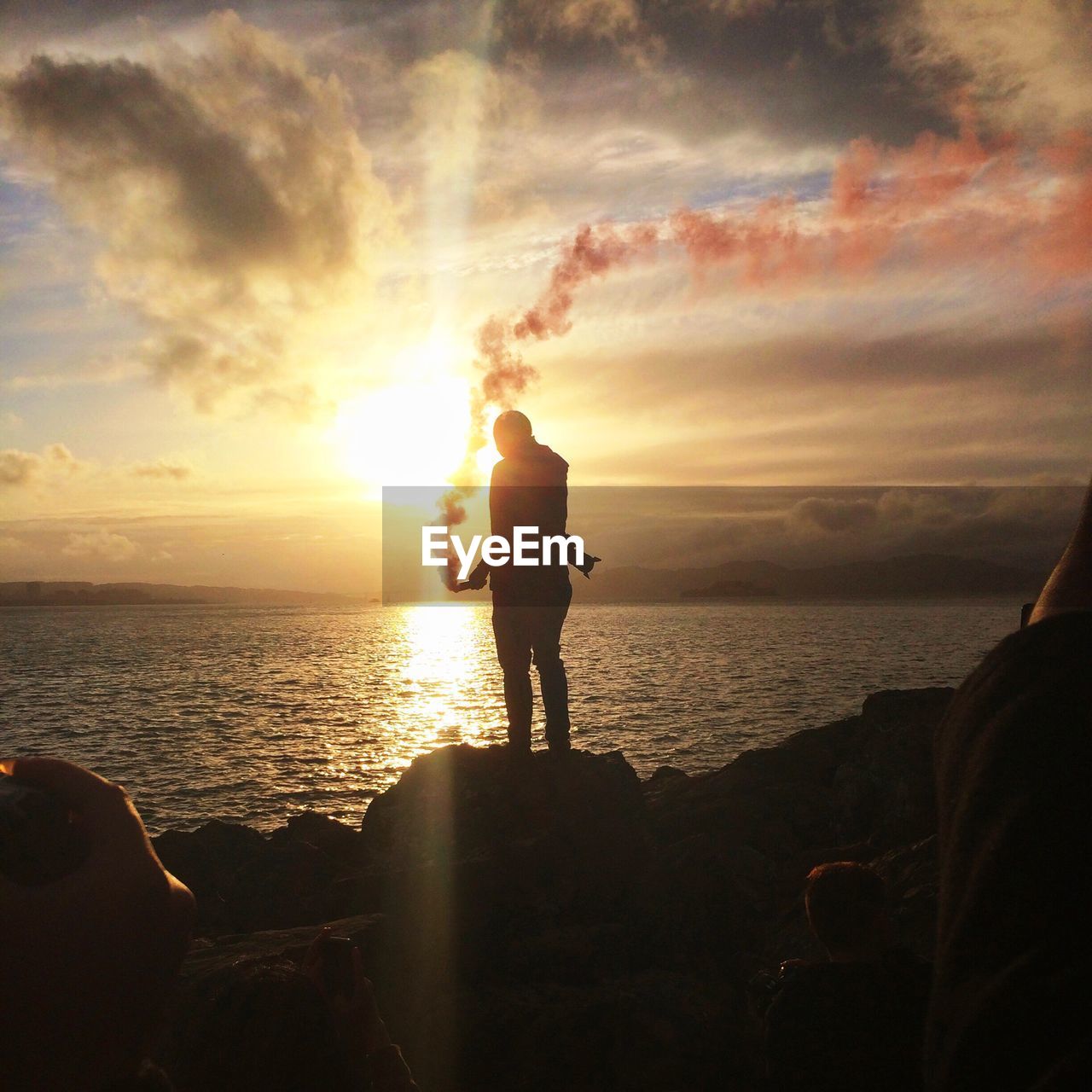 Man holding distress flare while standing on rock against sea during sunset