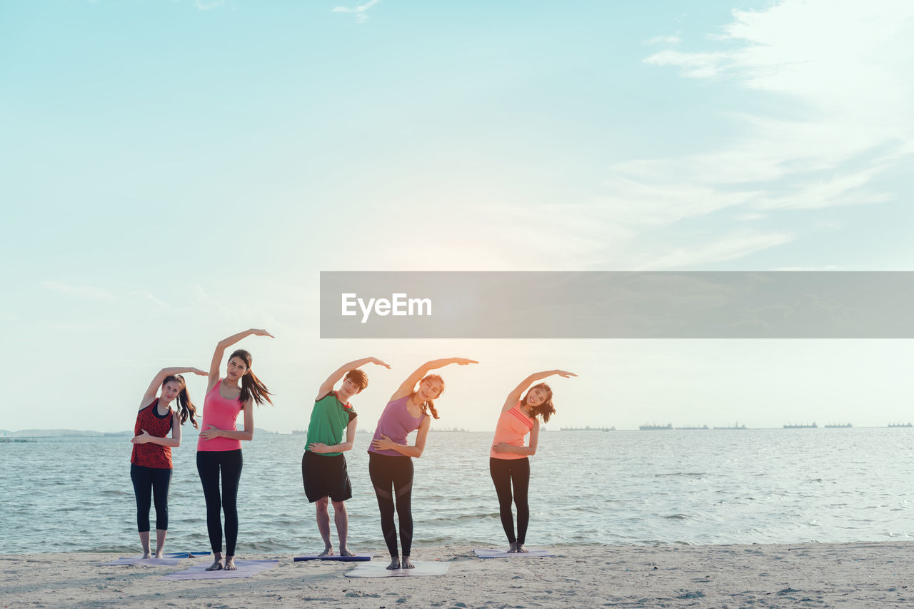 Young friends stretching arms while standing on shore at beach during sunny day