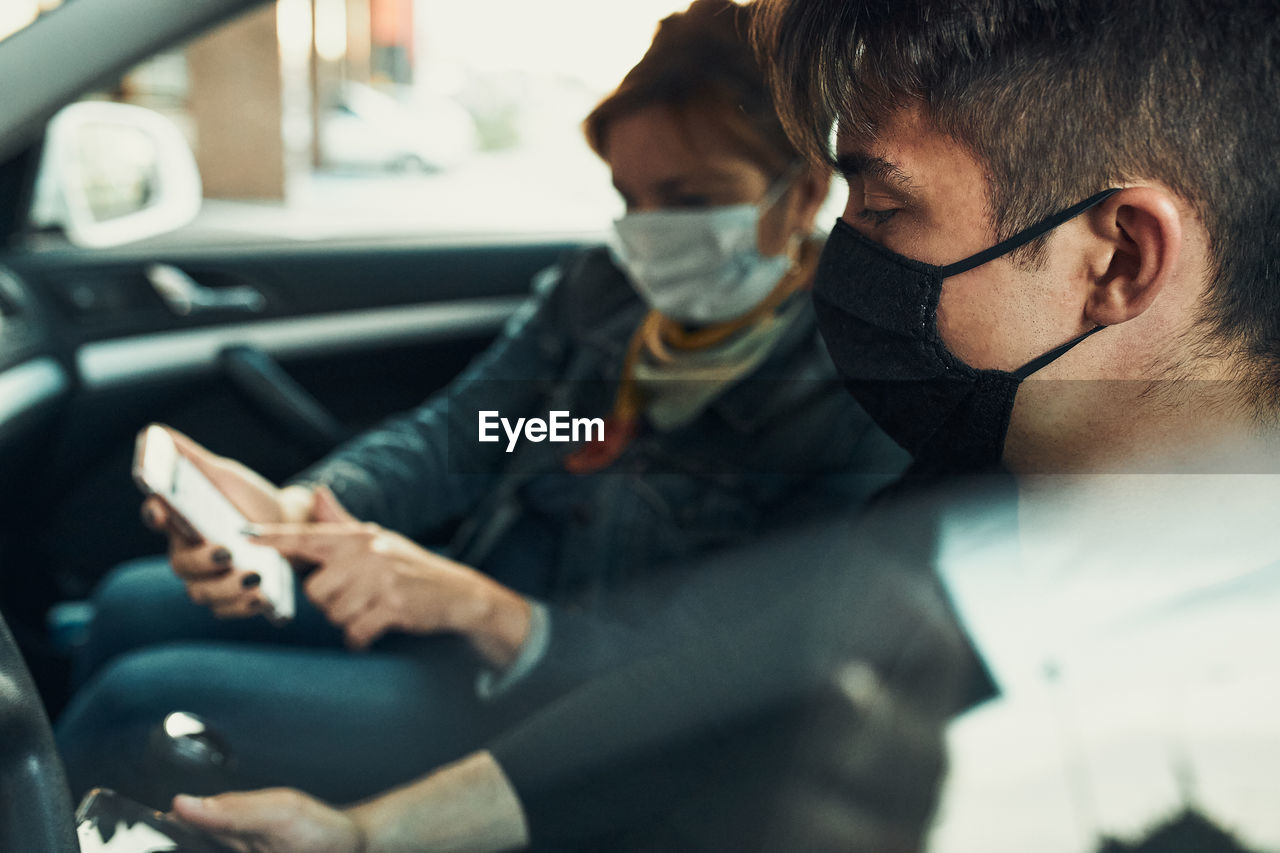 Man and woman sitting in a car using smartphones wearing the face masks to avoid virus infection