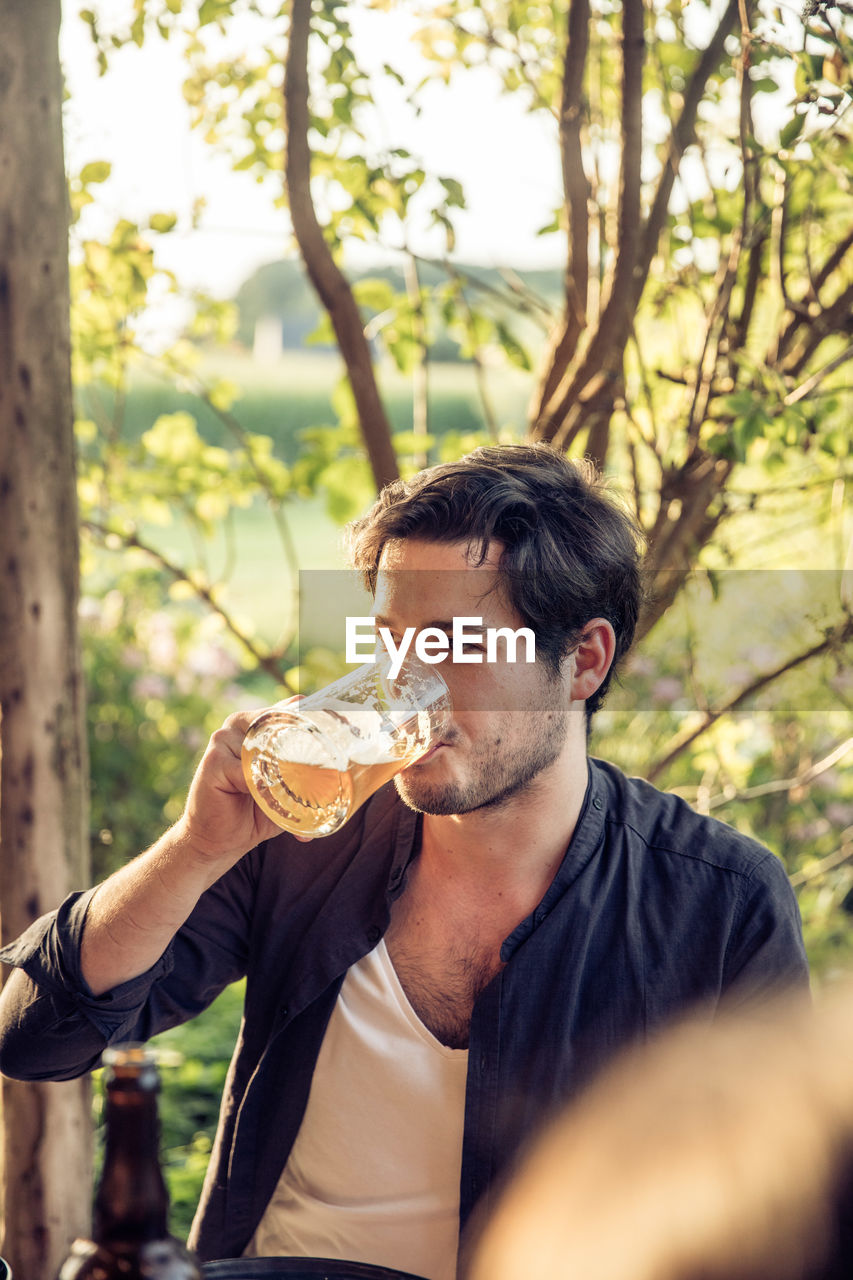 Young man looking away while drinking beer in glass against tree