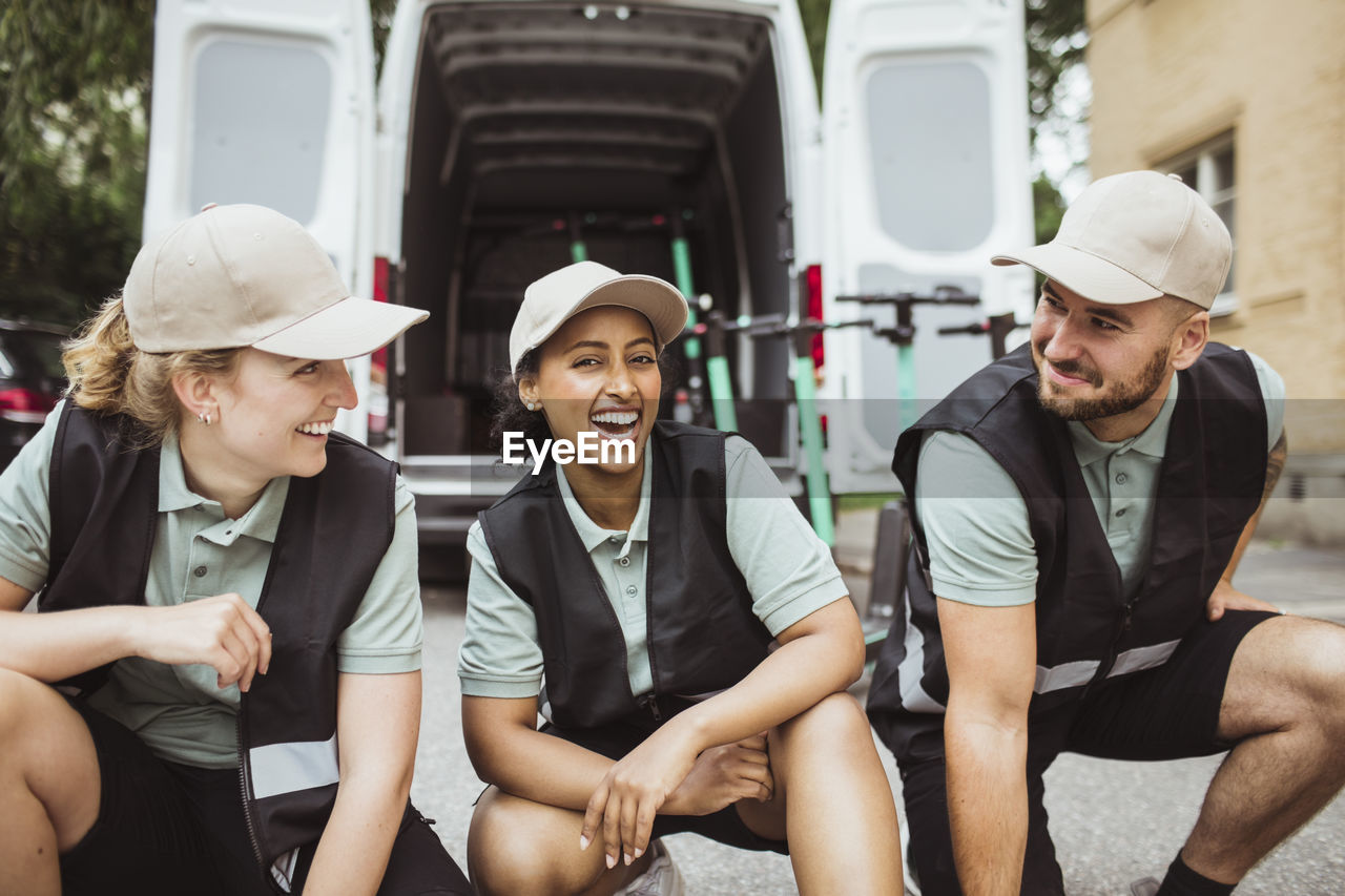 Portrait of female worker laughing while crouching amidst coworkers looking at each other against delivery van