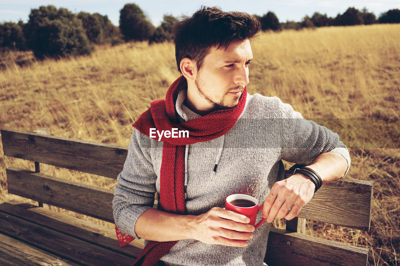 YOUNG MAN DRINKING COFFEE WHILE SITTING ON SEAT AT BEACH