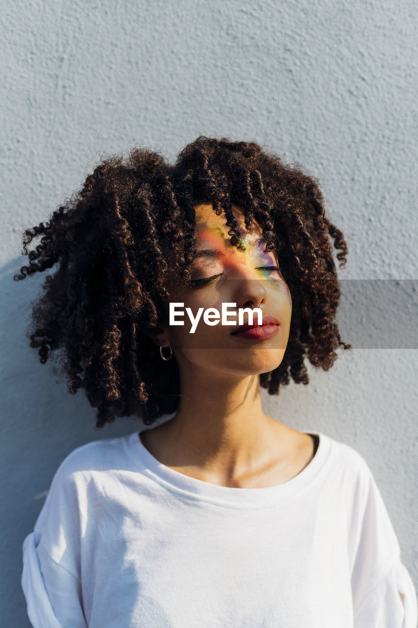 Young woman with eyes closed enjoying rainbow sunbeam in front of wall