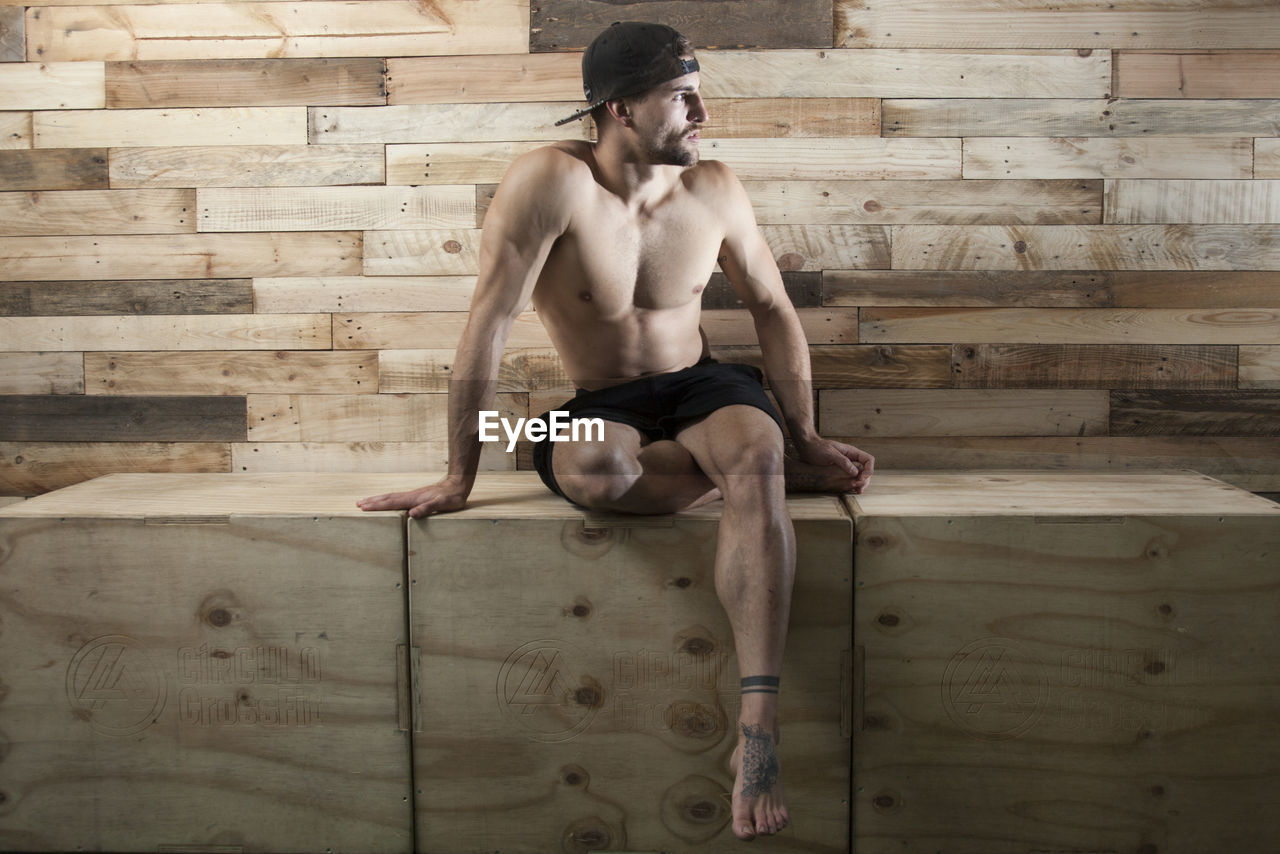 Full length of shirtless man sitting on wood